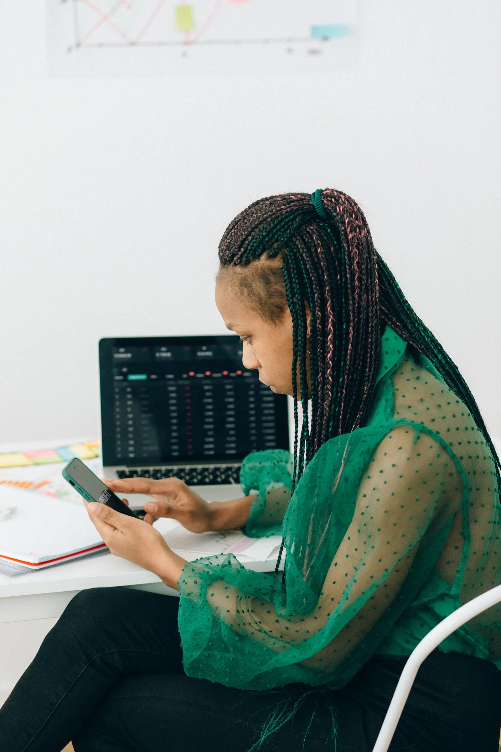A woman checks market information on her phone beside a laptop.